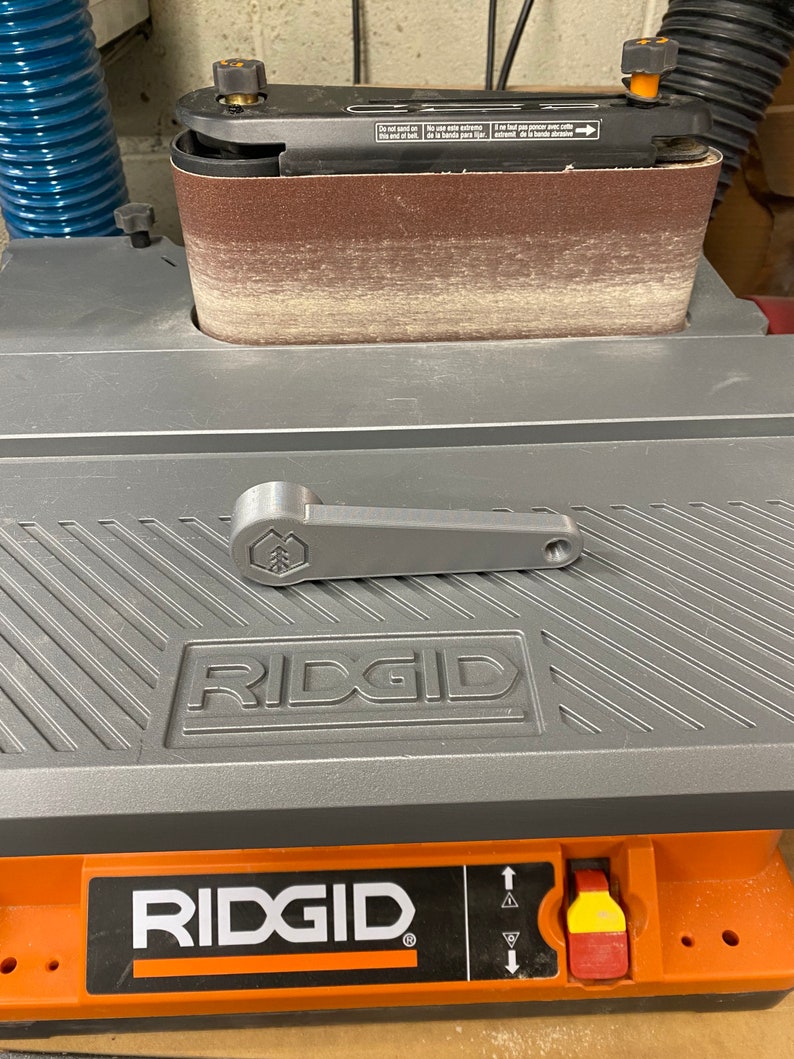 May include: A gray and orange Ridgid belt sander with a gray lever handle. The sander has a brown sanding belt and a black and orange dust collection port. The sander is on a gray surface with a black and white striped pattern.