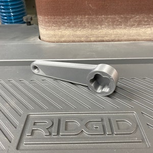 May include: A gray 3D printed wrench with a six-point socket. The wrench is lying on a gray surface with the word "RIDGID" embossed on it.