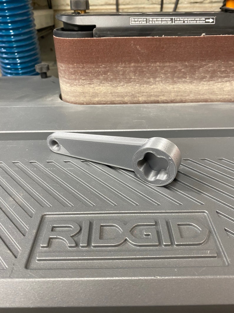 May include: A gray 3D printed wrench with a six-point socket. The wrench is lying on a gray surface with the word "RIDGID" embossed on it.