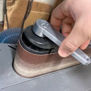 May include: A close-up of a gray 3D printed sanding belt tensioner knob on a belt sander. The knob has a textured surface and a hexagonal shape. The sander has a brown sanding belt.