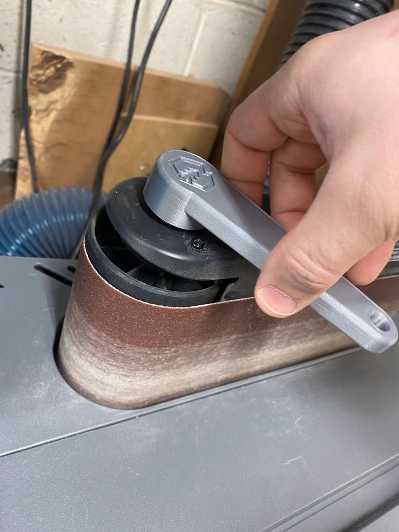 May include: A close-up of a gray 3D printed sanding belt tensioner knob on a belt sander. The knob has a textured surface and a hexagonal shape. The sander has a brown sanding belt.