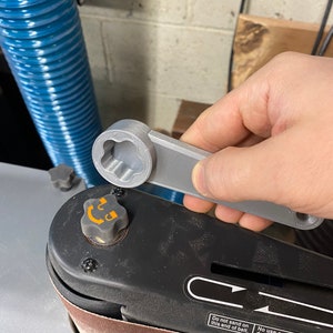 May include: A gray 3D printed wrench with a star-shaped head is being used to tighten a knob on a black sanding machine. The machine has a brown sanding belt and a label that reads "Do not sand on this end of belt. No use este extremo de la banda para lijar."