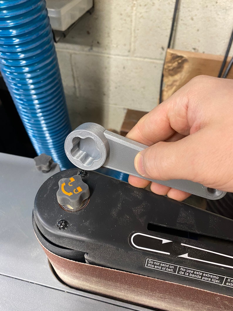 May include: A gray 3D printed wrench with a star-shaped head is being used to tighten a knob on a black sanding machine. The machine has a brown sanding belt and a label that reads "Do not sand on this end of belt. No use este extremo de la banda para lijar."