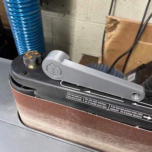 May include: A close-up of a belt sander with a gray handle and a brown sanding belt. The sander has a warning label that reads "Do not sand on this end of belt. No use este extremo de la banda para lijar. Il ne faut pas poncer avec cette extremit de la bande abrasive."