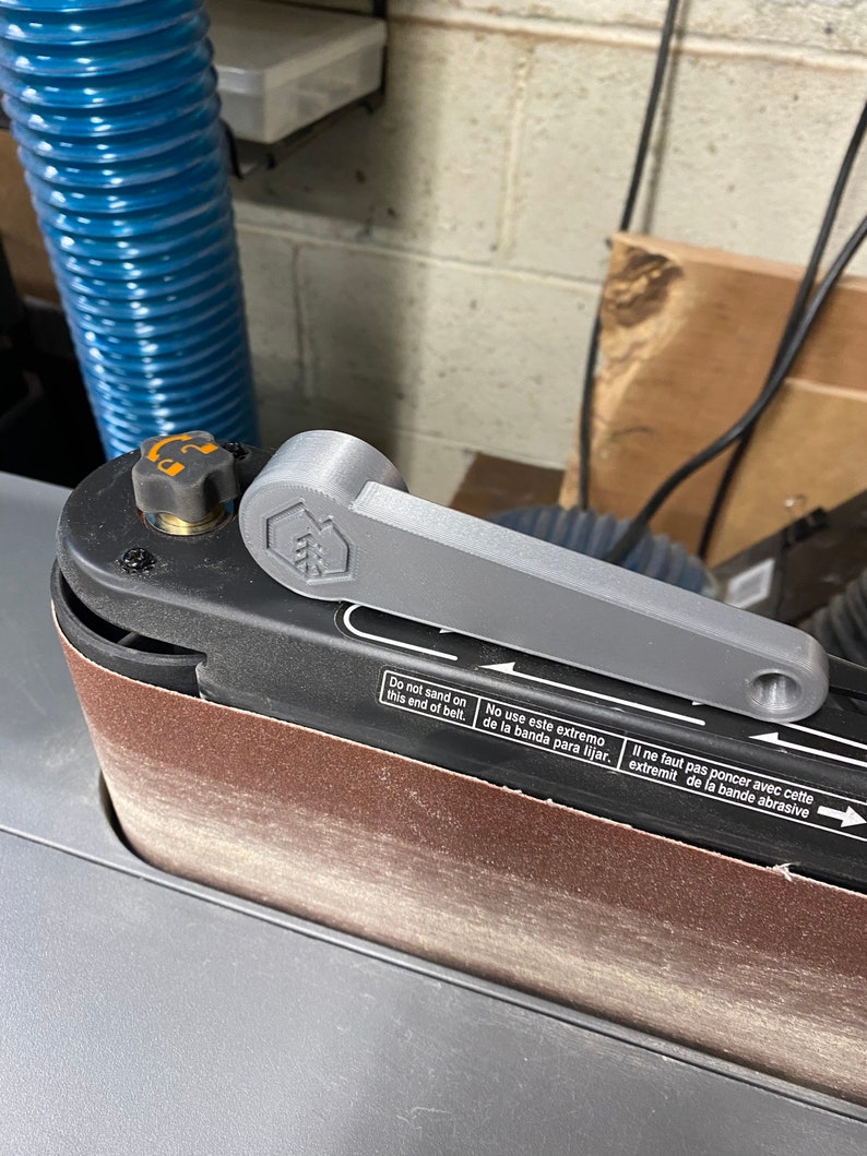 May include: A close-up of a belt sander with a gray handle and a brown sanding belt. The sander has a warning label that reads "Do not sand on this end of belt. No use este extremo de la banda para lijar. Il ne faut pas poncer avec cette extremit de la bande abrasive."