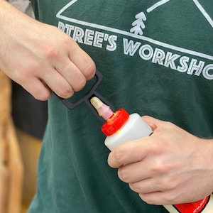 May include: A person is using a black plastic tool to apply glue from a white bottle with a red cap. The bottle is labeled "NET WT. 16 OZ (1 LB)". The person is wearing a green shirt with the text "TREE'S WORKSHOP" and a white pine tree graphic.