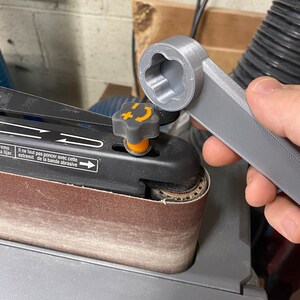 May include: A gray 3D printed wrench with a star-shaped opening is being used to tighten a knob on a belt sander. The sander has a brown sanding belt and a black and orange knob.