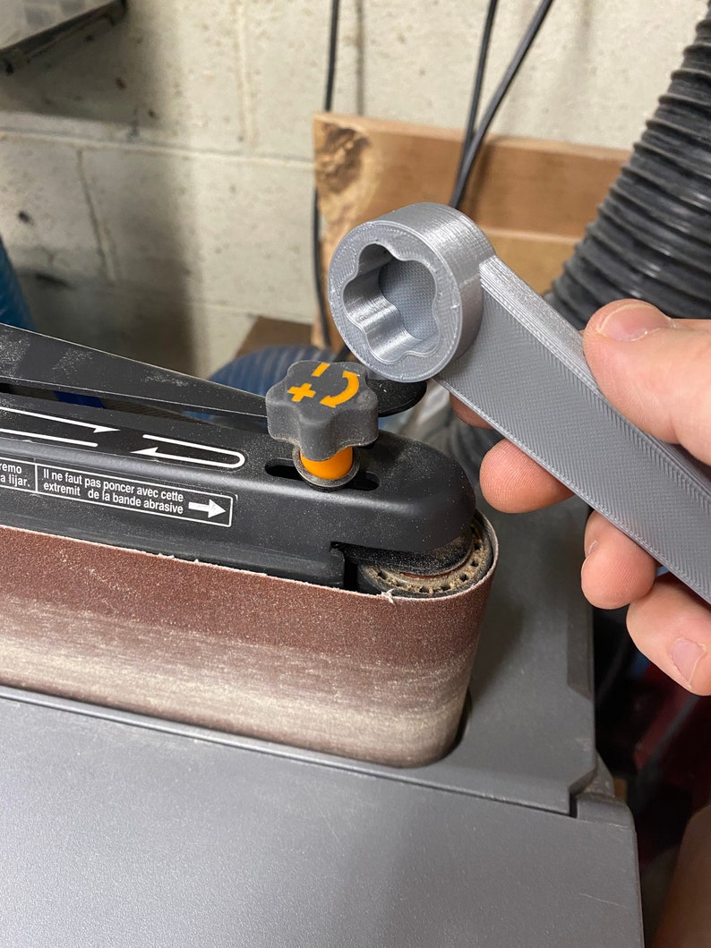 May include: A gray 3D printed wrench with a star-shaped opening is being used to tighten a knob on a belt sander. The sander has a brown sanding belt and a black and orange knob.