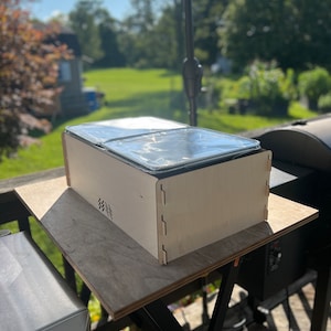 May include: A wooden box with a lid, containing two metal baking pans. The box is sitting on a wooden table, with a black grill in the background.
