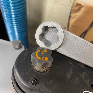 May include: A gray 3D printed wrench with a flower-shaped opening, resting on top of a black metal object with a gray knob. The knob has a yellow symbol with a curved line and a letter "D".