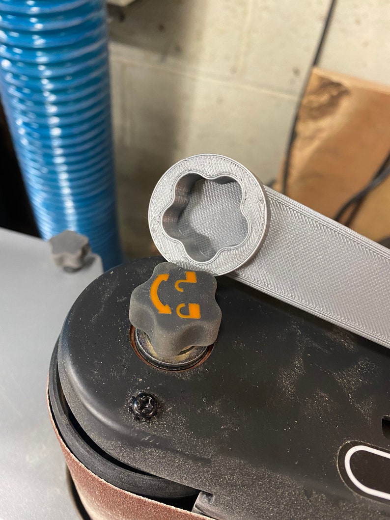 May include: A gray 3D printed wrench with a flower-shaped opening, resting on top of a black metal object with a gray knob. The knob has a yellow symbol with a curved line and a letter "D".