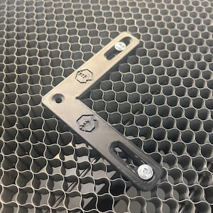 Puede incluir: Un soporte en forma de L de plástico negro con dos agujeros para tornillos y un logotipo en cada lado. El soporte está sobre una superficie de panal gris.