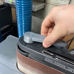 May include: A close-up of a gray belt sander with a black sanding belt. The sander has a gray lever that is being turned by a hand. The sander has a warning label that reads "Do not sand on this end of belt. No use este extremo de la banda para lijar. Il ne faut pas utiliser cette extr&eacute;mit&eacute; de la bande pour poncer."