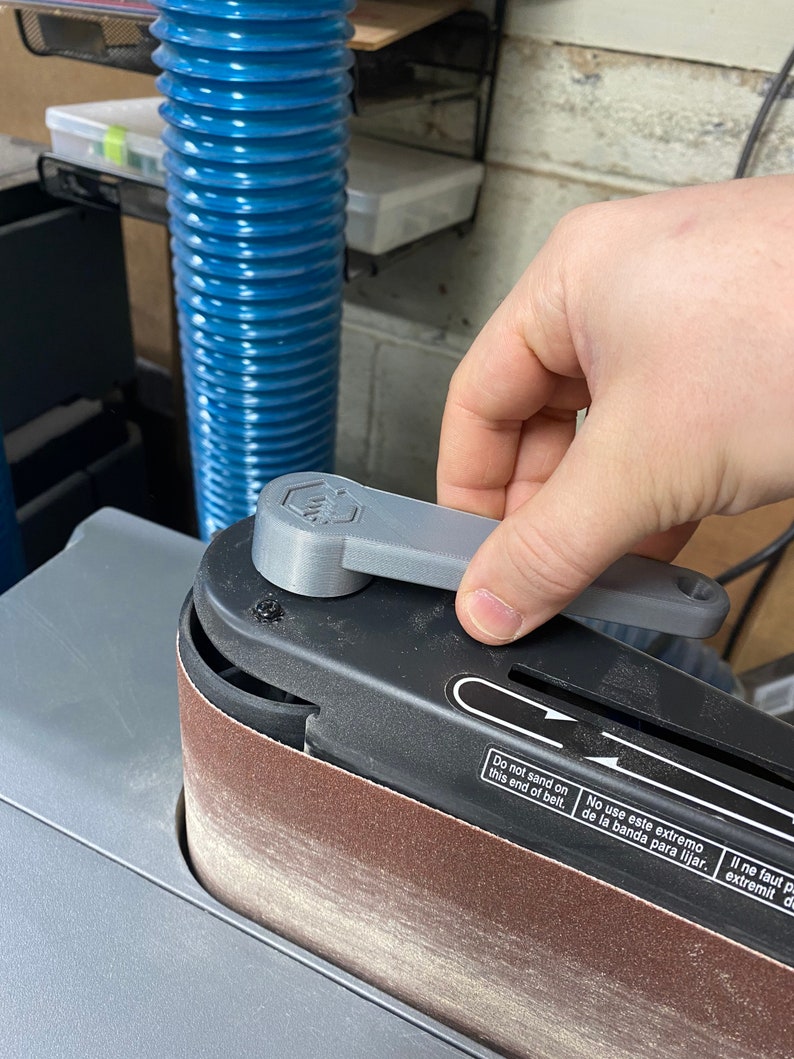 May include: A close-up of a gray belt sander with a black sanding belt. The sander has a gray lever that is being turned by a hand. The sander has a warning label that reads "Do not sand on this end of belt. No use este extremo de la banda para lijar. Il ne faut pas utiliser cette extr&eacute;mit&eacute; de la bande pour poncer."
