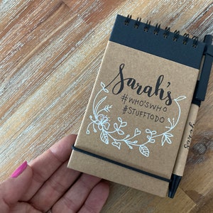 May include: A brown notebook with a black elastic band and a black pen. The notebook has a floral design and the text "Sarah's #WHOSWHO #STUFFTODO" written on it.