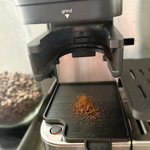 Op de afbeelding: Een roestvrijstalen koffiemolen met een zwart kunststof doseergedeelte voorzien van het woord grind en een pijl naar beneden. Een klein hoopje donkerbruine gemalen koffie ligt op de zwarte rechthoekige lekbak onder de uitloop.