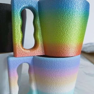 May include: Two stacked, rainbow-coloured mugs with a textured surface. The mugs have a unique handle design with a large opening. The colours transition from blue to green, orange, and pink. A spoon rests on top of the upper mug.