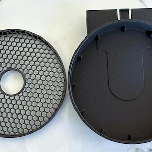 Puede incluir: Componentes de altavoz de plástico negro. La imagen muestra una rejilla circular con un patrón de panal y una abertura redonda en el centro. Junto a ella, una carcasa de altavoz negra y redonda con un recorte en forma de U y un accesorio rectangular.