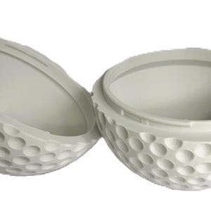 Könnte beinhalten: Ein weißer, kugelförmiger Behälter in Form eines Golfballs, der in zwei offene Hälften geteilt ist. Die Außenfläche weist die charakteristische genoppte Textur eines Golfballs auf, während das Innere glatt und hohl für Aufbewahrungszwecke ist.