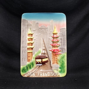 May include: A vintage ceramic tile depicting a San Francisco cable car traveling up a hill between two buildings with pagoda-style roofs. The tile is painted in shades of brown, red, yellow, and green, and the text "SAN FRANCISCO" is printed in white at the bottom.