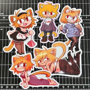 Peut inclure: Plusieurs autocollants colorés représentant un personnage de chat de dessin animé dans différentes poses et tenues. Un autocollant porte le mot "GAY". Les autocollants sont sur un tapis de coupe noir avec des mesures en centimètres et en pouces.