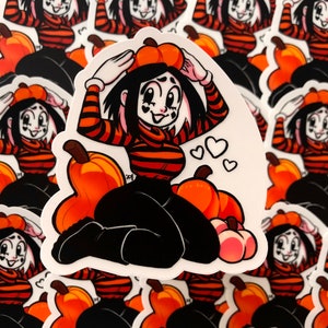 Puede incluir: Una pegatina con un personaje de dibujos animados que lleva una camisa a rayas y un sombrero de calabaza. El personaje está rodeado de calabazas y corazones.