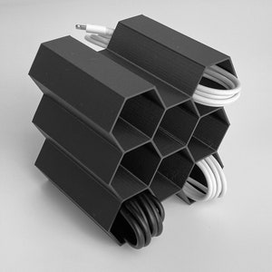 May include: A black hexagonal cable organizer with multiple compartments for storing and organizing cords and cables. The organizer is filled with white and black cables.