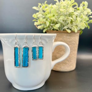 May include: Three pairs of rectangular, turquoise-colored earrings with silver-colored metal frames and hooks. The earrings are displayed inside a white teacup. A small potted plant is in the background.