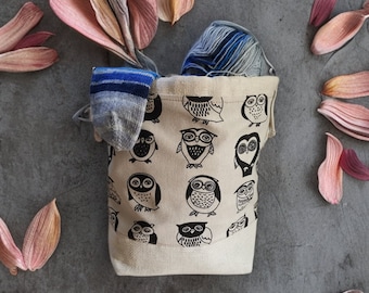 Puede incluir: Una bolsa de lona beige con estampado de búho en blanco y negro. La bolsa está llena de hilo y un calcetín azul.