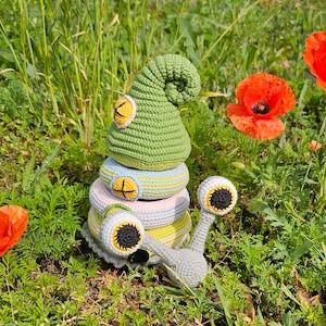 Puede incluir: Un juguete de caracol de crochet con una concha verde, ojos amarillos y un cuerpo gris. El caracol está sentado en una cama de hierba verde con amapolas rojas.