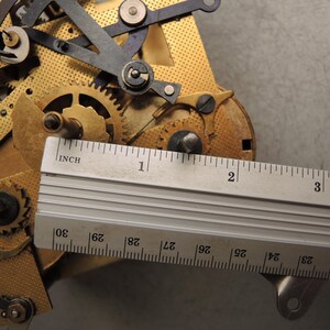 May include: Close-up of a clock mechanism with brass gears and metal components. A ruler is placed over the mechanism, showing measurements in inches. The image highlights the intricate details of the clock's internal workings.
