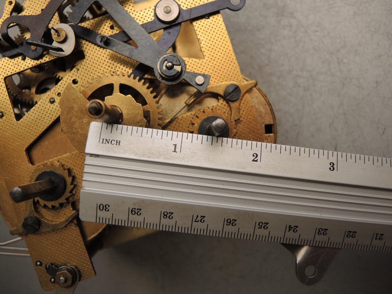May include: Close-up of a clock mechanism with brass gears and metal components. A ruler is placed over the mechanism, showing measurements in inches. The image highlights the intricate details of the clock's internal workings.