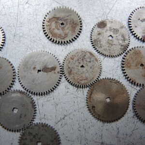 May include: A collection of small, circular metal gears with toothed edges. The gears have a square hole in the center and show signs of wear and rust, with a silver and brown color palette. These components are likely from a mechanical device.
