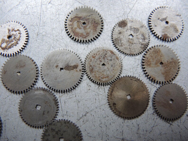 May include: A collection of small, circular metal gears with toothed edges. The gears have a square hole in the center and show signs of wear and rust, with a silver and brown color palette. These components are likely from a mechanical device.