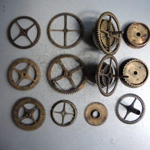May include: A collection of various antique clock gears in shades of gold and bronze, arranged on a light gray surface. The gears vary in size and design, showcasing intricate details and a vintage aesthetic.