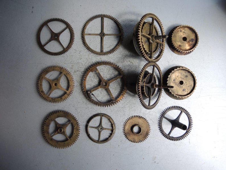 May include: A collection of various antique clock gears in shades of gold and bronze, arranged on a light gray surface. The gears vary in size and design, showcasing intricate details and a vintage aesthetic.