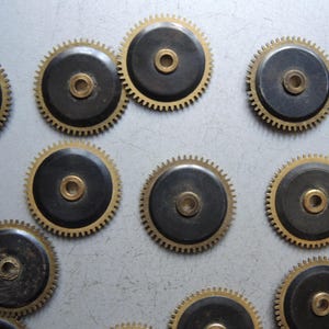 May include: Close-up of several watch gears. Each gear has a black center with a gold-colored center and a gold-colored toothed outer ring. The gears are arranged in a scattered pattern.