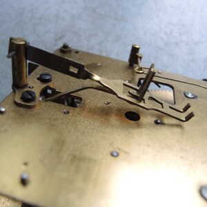 May include: Close-up of a brass clock mechanism. The image shows the intricate details of the clock's internal components, including gears, levers, and screws. The brass has a warm, golden hue, and the parts are arranged on a rectangular base.