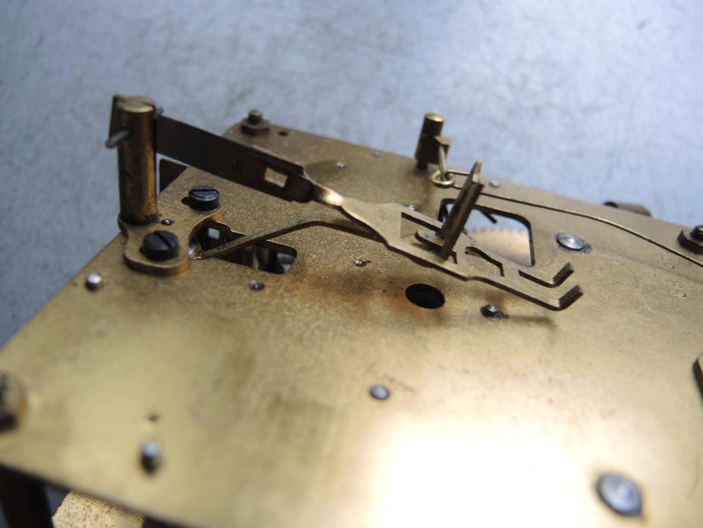 May include: Close-up of a brass clock mechanism. The image shows the intricate details of the clock's internal components, including gears, levers, and screws. The brass has a warm, golden hue, and the parts are arranged on a rectangular base.