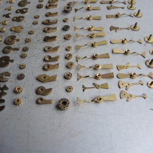 May include: A collection of small, vintage watch parts, including gears, levers, and other components. The parts are made of brass and are arranged on a gray surface. The image showcases a variety of shapes and sizes, ideal for watch repair or crafting.