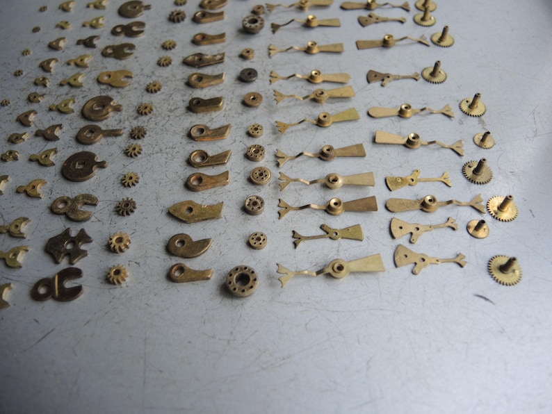 May include: A collection of small, vintage watch parts, including gears, levers, and other components. The parts are made of brass and are arranged on a gray surface. The image showcases a variety of shapes and sizes, ideal for watch repair or crafting.