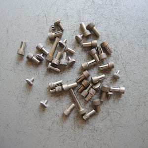 May include: A collection of small, silver-toned metal hardware components, each featuring a cylindrical body with a knurled, textured head. These fasteners vary in length and include slotted ends, appearing as specialized pins or adjustment screws.