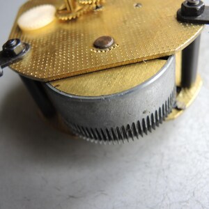 Op de afbeelding: Close-up van een uurwerk met messing en zilveren componenten. De messing plaat heeft een getextureerd oppervlak met kleine gaatjes. Een zilveren tandwiel is zichtbaar, samen met zwarte metalen steunen en schroeven. Het uurwerk is waarschijnlijk voor reparatie of restauratie.