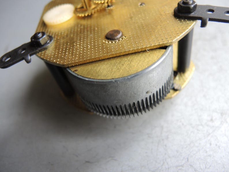 Op de afbeelding: Close-up van een uurwerk met messing en zilveren componenten. De messing plaat heeft een getextureerd oppervlak met kleine gaatjes. Een zilveren tandwiel is zichtbaar, samen met zwarte metalen steunen en schroeven. Het uurwerk is waarschijnlijk voor reparatie of restauratie.