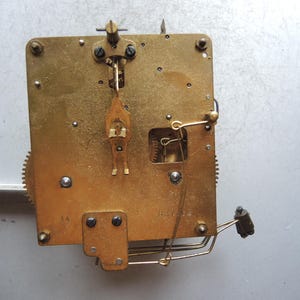 May include: Close-up of a vintage clock movement. The brass mechanism features gears, levers, and screws, with a rectangular base. The intricate design showcases the internal workings of a clock, highlighting its mechanical components.
