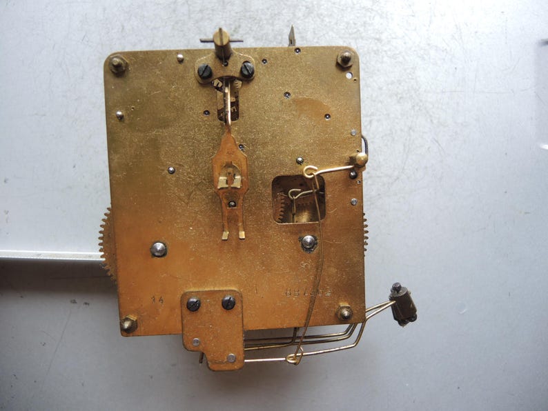 May include: Close-up of a vintage clock movement. The brass mechanism features gears, levers, and screws, with a rectangular base. The intricate design showcases the internal workings of a clock, highlighting its mechanical components.