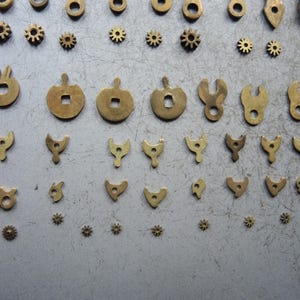 May include: An assortment of small, antique brass watch parts, including gears, levers, and decorative shapes. The components are arranged on a gray surface, showcasing their intricate designs and varying sizes. These parts are likely for watch repair or restoration.
