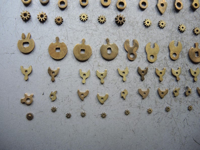 May include: An assortment of small, antique brass watch parts, including gears, levers, and decorative shapes. The components are arranged on a gray surface, showcasing their intricate designs and varying sizes. These parts are likely for watch repair or restoration.