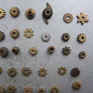 May include: A selection of small, brass-coloured gears in various sizes and shapes, including circular, star-shaped, and square-holed designs. The gears are scattered on a grey surface, showcasing their intricate details.