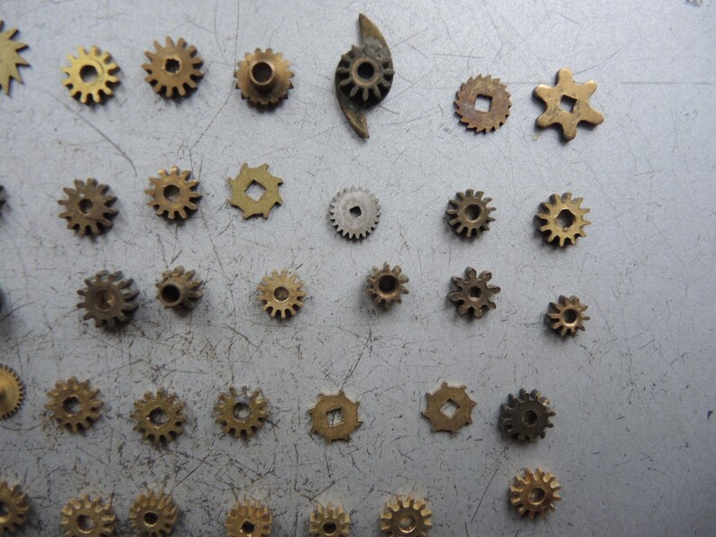 May include: A selection of small, brass-coloured gears in various sizes and shapes, including circular, star-shaped, and square-holed designs. The gears are scattered on a grey surface, showcasing their intricate details.